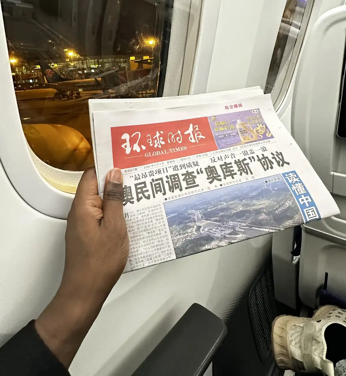 Andrew Miracle holds a Chinese-language Global Times newspaper by an airplane window at night